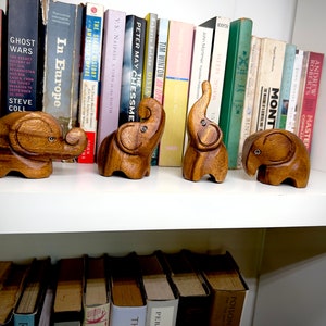 May include: Five wooden elephant figurines on a white shelf with books behind them. The elephants are all different sizes and have a natural wood finish.