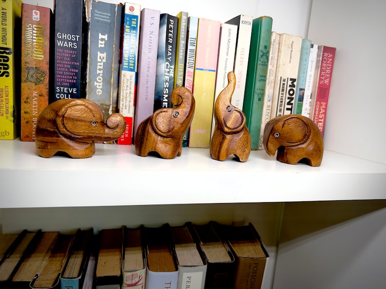 May include: Five wooden elephant figurines on a white shelf with books behind them. The elephants are all different sizes and have a natural wood finish.