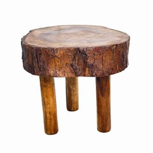Peut inclure: Un petit tabouret rond en bois avec un dessus en tronc d'arbre naturel et trois pieds robustes. Le tabouret est en bois brun clair, avec le dessus montrant le grain et la texture naturels du bois. Les pieds sont d'une teinte plus foncée de brun.