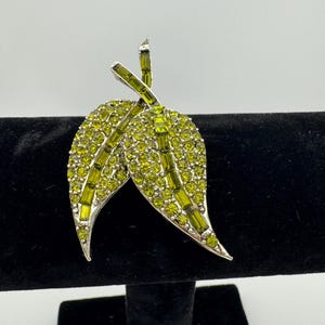 May include: A silver tone brooch featuring two green leaves crafted from rhinestones. The leaves are adorned with clear and green rhinestones.