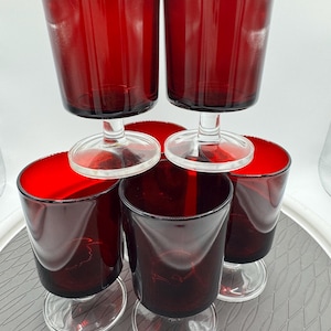 May include: A set of five ruby red glass goblets with clear glass stems and circular bases. The deep red bowls feature a smooth, cylindrical shape with a slightly flared rim, providing a consistent look across all five drinking vessels.