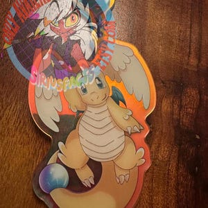 Puede incluir: Un colgante acrílico transparente con un dragón de dibujos animados con un cuerpo naranja claro, alas blancas y un orbe azul y morado. El colgante tiene un cordón azul y amarillo y un segundo colgante con un personaje de Hazbin Hotel. El texto en el colgante dice "ONLY AVAILABLE AT ETSY.COM/SIRIUSFAIRY".