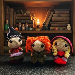 Pode incluir: Três bonecas de crochê alinhadas. A boneca da esquerda tem cabelo preto, um top vermelho e uma saia roxa. A do meio tem cabelo laranja encaracolado e um casaco verde. A da direita tem cabelo loiro, um capuz bordô e um vestido vermelho.
