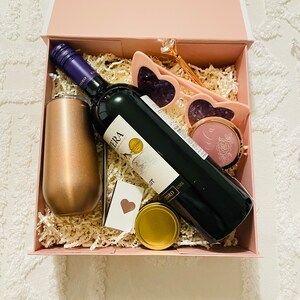 May include: A pink gift box filled with a bottle of red wine, a gold tumbler, a pair of pink heart-shaped sunglasses, a small round mirror with a floral design, and a gold lidded jar.