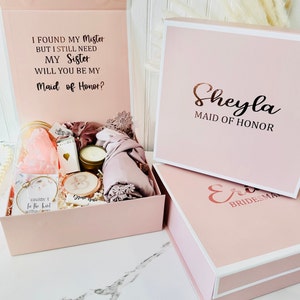 May include: Pink gift box with a white lid and a white border. The lid has the text "I found my Mister but I still need my Sister. Will you be my Maid of Honor?" The box has the text "Sheyla Maid of Honor" on the lid. The box is open and contains a variety of items, including a candle, a scarf, and a small jar.