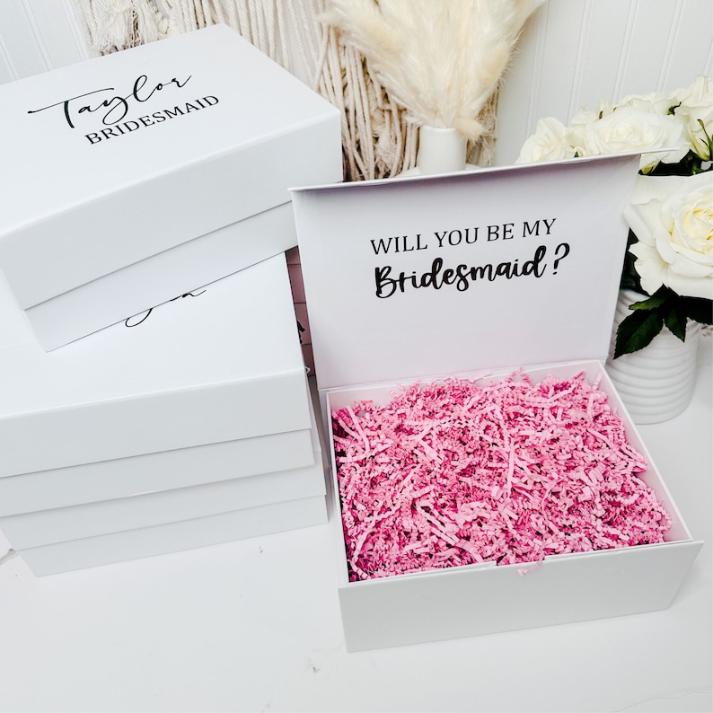 May include: White gift box with a pink tissue paper filler. The lid of the box has the text "Will you be my Bridesmaid?" printed in black.