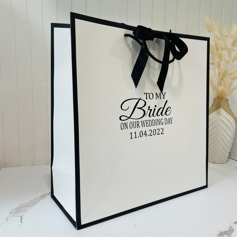 K&ouml;nnte beinhalten: Eine wei&szlig;e Geschenkt&uuml;te mit schwarzem Rand und schwarzem Band. Auf der T&uuml;te steht der Text "To My Bride On Our Wedding Day 11.04.2022".