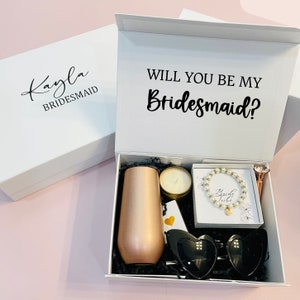 May include: White gift box with a black lid that says "Will you be my Bridesmaid?" The box is open and contains a rose gold tumbler, a candle, a bracelet with a charm that says "Bride Tribe", a pair of black heart-shaped sunglasses, and a gold pen with a diamond-shaped top.