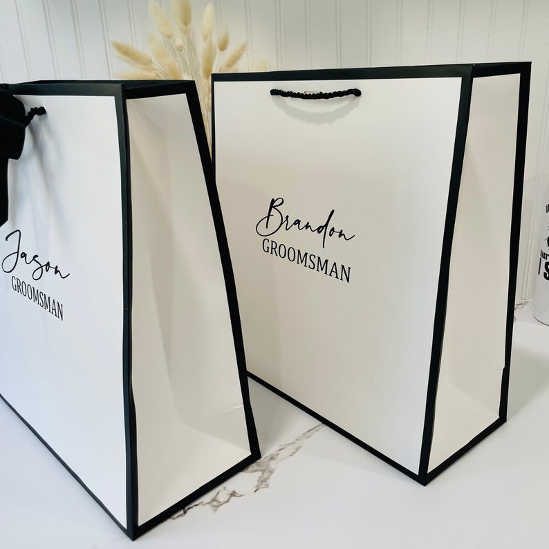 K&ouml;nnte beinhalten: Zwei wei&szlig;e Geschenkt&uuml;ten mit schwarzen Henkeln und schwarzem Besatz. Die Taschen haben die Namen "Jason" und "Brandon" mit dem Wort "Groomsman" darunter aufgedruckt.