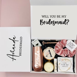 May include: A white gift box with a pink and gold bridesmaid gift set. The box has the text "Will you be my Bridesmaid?" on the lid. The gift set includes a tumbler, a candle, a hair scrunchie, a small mirror, a bracelet, and a lip balm.