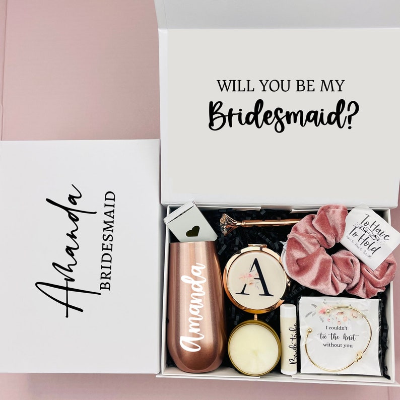 May include: A white gift box with a pink and gold bridesmaid gift set. The box has the text "Will you be my Bridesmaid?" on the lid. The gift set includes a tumbler, a candle, a hair scrunchie, a small mirror, a bracelet, and a lip balm.