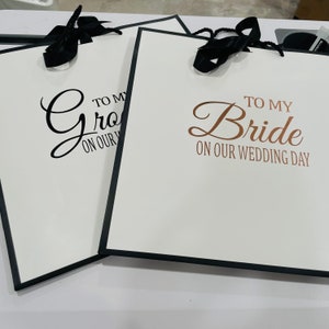 K&ouml;nnte beinhalten: Zwei wei&szlig;e Geschenkt&uuml;ten mit schwarzen Schleifenb&auml;ndern. Die Tasche links sagt "To my Groom on our Wedding Day" in schwarzen Buchstaben. Die Tasche rechts sagt "To my Bride on our Wedding Day" in schwarzen Buchstaben mit "Bride" in goldenen Buchstaben.