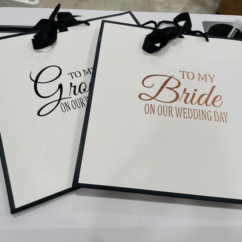 K&ouml;nnte beinhalten: Zwei wei&szlig;e Geschenkt&uuml;ten mit schwarzen Schleifenb&auml;ndern. Die Tasche links sagt "To my Groom on our Wedding Day" in schwarzen Buchstaben. Die Tasche rechts sagt "To my Bride on our Wedding Day" in schwarzen Buchstaben mit "Bride" in goldenen Buchstaben.