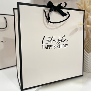 K&ouml;nnte beinhalten: Wei&szlig;e Geschenkt&uuml;te mit schwarzem Besatz und schwarzem Band. Die Tasche hat den Text "Latasha Happy Birthday" auf der Vorderseite.