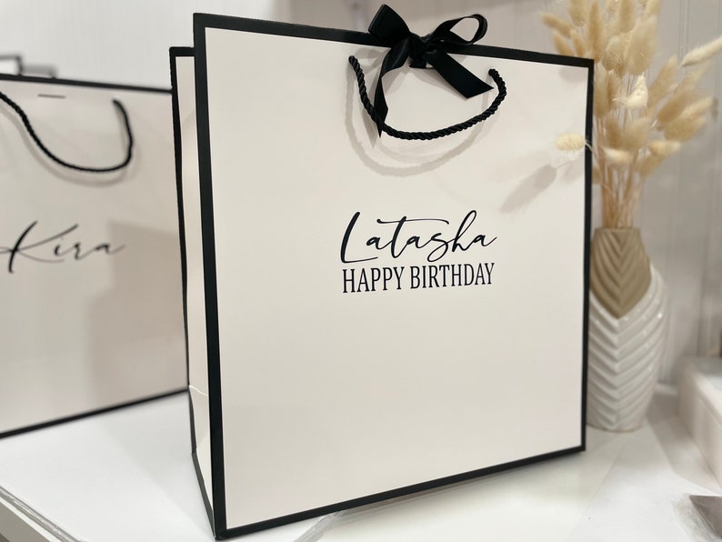 K&ouml;nnte beinhalten: Wei&szlig;e Geschenkt&uuml;te mit schwarzem Besatz und schwarzem Band. Die Tasche hat den Text "Latasha Happy Birthday" auf der Vorderseite.