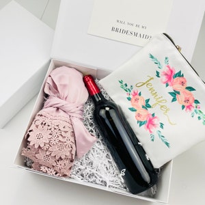 May include: A white gift box with a pink satin robe, a bottle of red wine, and a white cosmetic bag with floral print and the name "Jennifer" in gold lettering. The box also includes a card that says "Will you be my bridesmaid?"