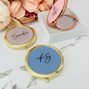 May include: Two gold compact mirrors with personalized names. One mirror is pink with the name "Sandra" and the other is blue with the initials "Ky".