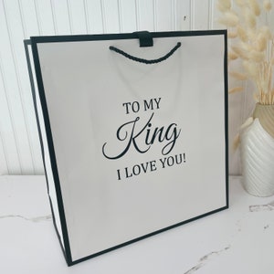 K&ouml;nnte beinhalten: Wei&szlig;e Geschenkt&uuml;te mit schwarzem Besatz und Kordelgriffen. Die Tasche tr&auml;gt den Aufdruck "To My King I Love You!" auf der Vorderseite.