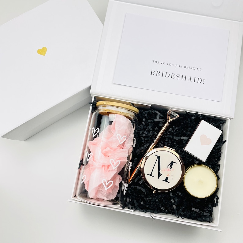 May include: A white gift box with a black and white patterned tissue paper, a clear glass jar with a pink and white heart design, a gold-colored compact mirror with the letter "M" on it, a gold-colored stick with a diamond-shaped top, a small white candle, and a white card with the text "Thank you for being my bridesmaid!"