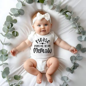 May include: A white baby onesie with black text that reads "Please pass me to my Memaw". A baby is wearing the onesie and has a white bow in their hair.
