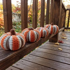 Puede incluir: Cuatro calabazas tejidas a mano dispuestas en fila sobre una barandilla de madera. Cada calabaza presenta rayas verticales alternas de hilo naranja quemado y gris claro, rematadas con un pequeño tallo marrón texturizado de ganchillo.