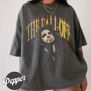 May include: A charcoal grey short-sleeve t-shirt featuring a graphic print of a man with dreadlocks. Above the portrait, the text THE FALL OFF is printed in a yellow and orange flame-like font. A red signature is visible to the right of the portrait.