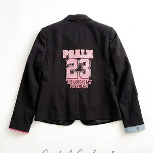 Puede incluir: Blazer negro con ribete rosa y pu&ntilde;o de mezclilla. La parte trasera del blazer presenta el texto "PSALM 23 THE LORD IS MY SHEPHERD" en letras rosas. El blazer es de estilo cl&aacute;sico con un corte entallado.