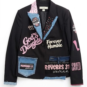 May include: Black blazer with denim accents and patches. Embellishments include pink sequined text that reads "God's Daughter", "Forever Humble", and "Saved". Denim pocket with "Proverbs 31 Woman" text. Pink high heel patch.