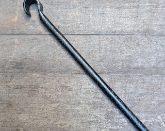 May include: A hand-forged black metal fire poker tool featuring a long, straight cylindrical shaft and a curved hook at the top end. The metal surface has a dark, textured finish consistent with ironwork, showing a simple and functional design.