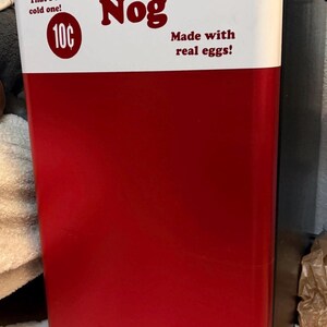 May include: A compact refrigerator with a red lower section and a white upper section. The white panel features the text DRINK Jugger-Nog, That's a cold one!, 10&cent;, and Made with real eggs! in dark red lettering. The sides of the unit are black.