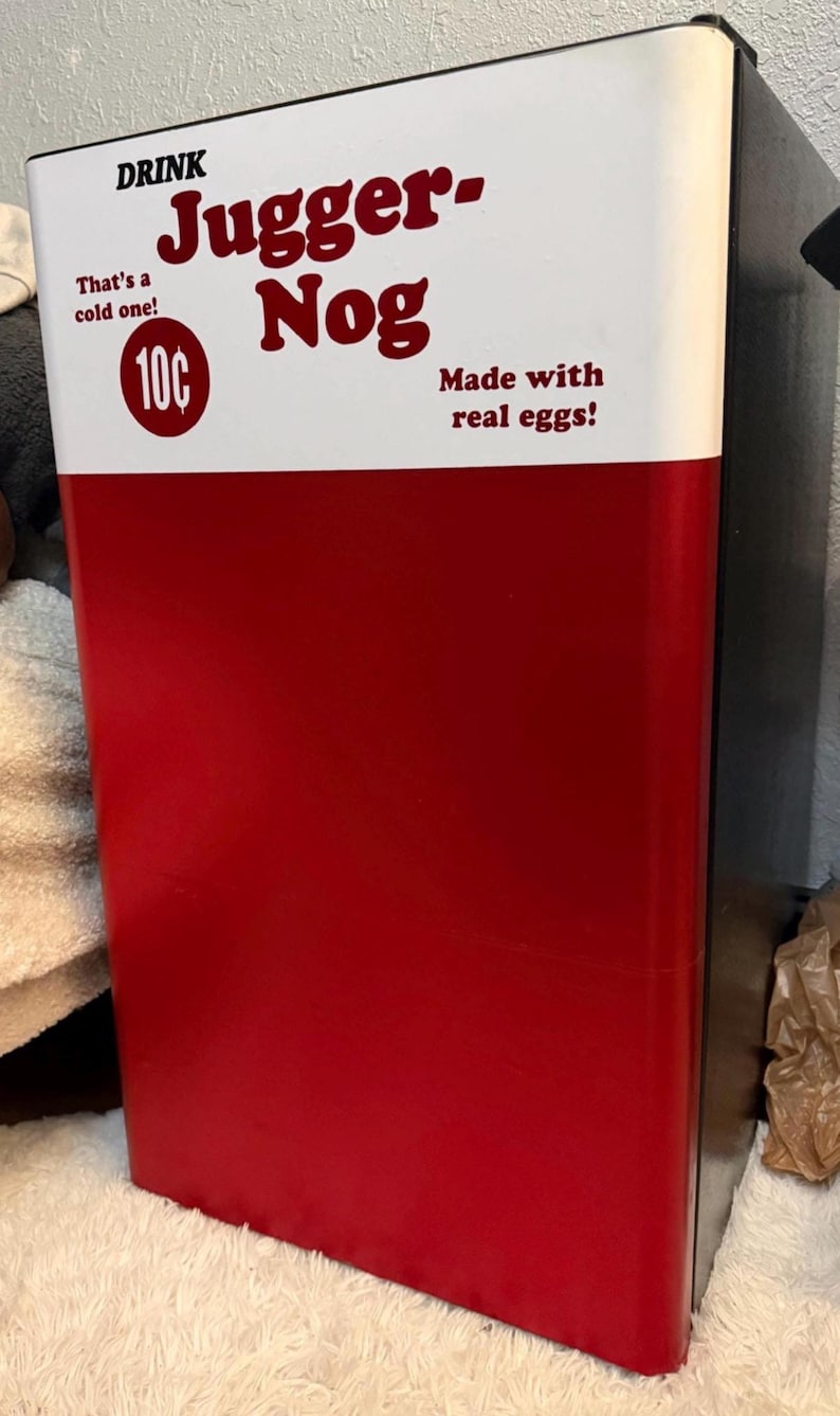 May include: A compact refrigerator with a red lower section and a white upper section. The white panel features the text DRINK Jugger-Nog, That's a cold one!, 10&cent;, and Made with real eggs! in dark red lettering. The sides of the unit are black.