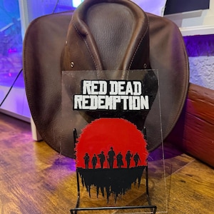 May include: A brown leather cowboy hat positioned behind a clear acrylic display stand. The stand features the white text RED DEAD REDEMPTION above a red circular graphic depicting the black silhouettes of seven figures standing in a line.