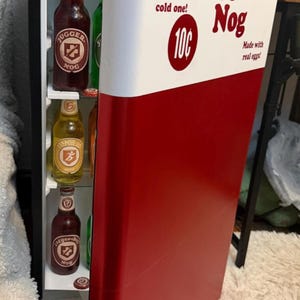 May include: A small refrigerator with a red and white door featuring the text Drink Jugger-Nog, That's a cold one!, 10c, and Made with real eggs!. Inside are glass shelves holding several glass bottles with labels including Jugger-Nog.