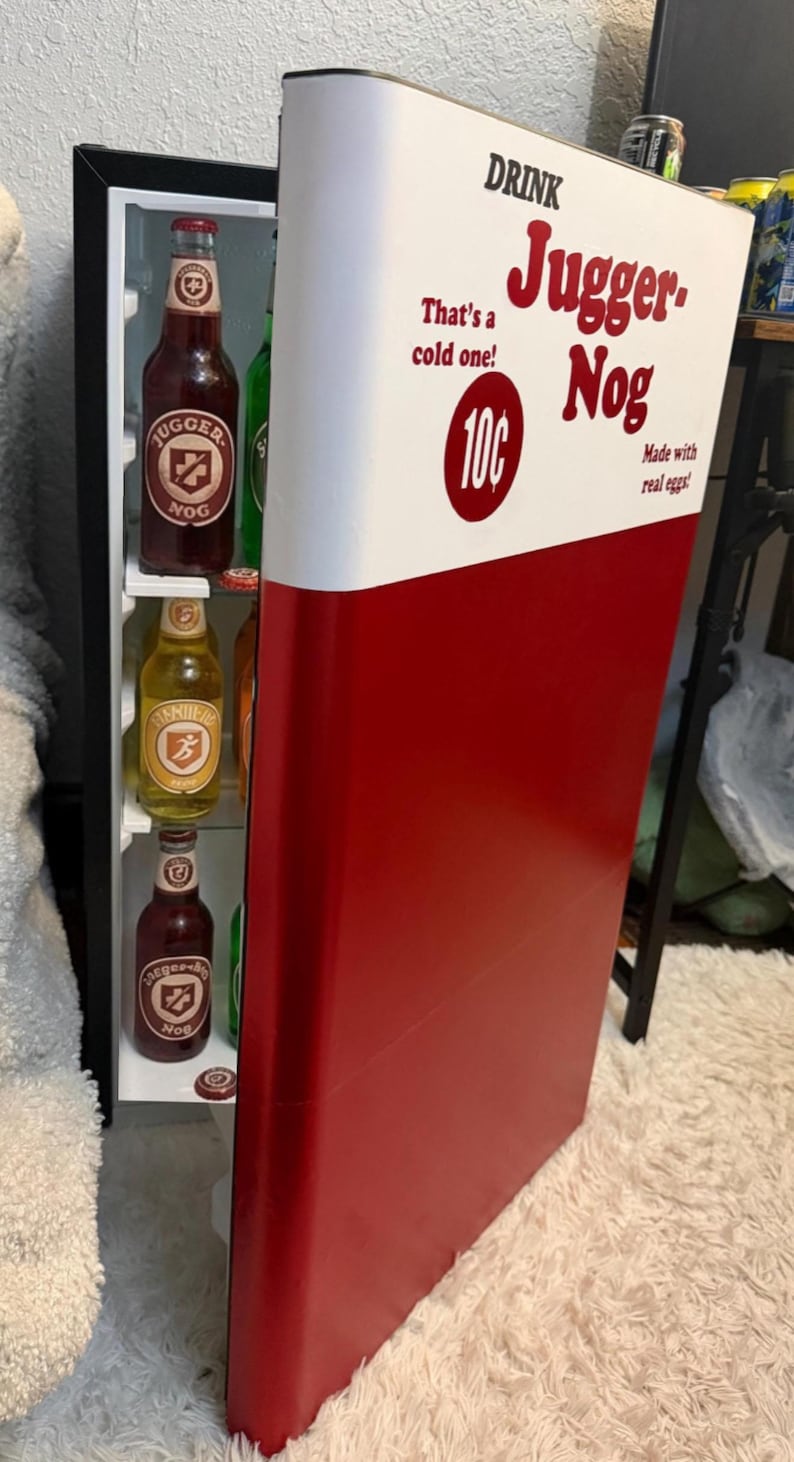 May include: A small refrigerator with a red and white door featuring the text Drink Jugger-Nog, That's a cold one!, 10c, and Made with real eggs!. Inside are glass shelves holding several glass bottles with labels including Jugger-Nog.