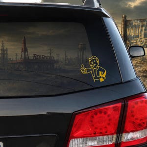 May include: A yellow vinyl decal of the Vault Boy character from the Fallout video game series, depicted giving a thumbs-up gesture. The decal is applied to the rear window of a black vehicle, positioned against a dark, post-apocalyptic landscape.