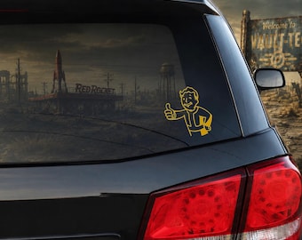 以下が含まれることがあります： ビデオゲームシリーズ「Fallout」のキャラクター、Vault Boyが親指を立てているポーズの黄色いビニール製デカール。黒い車のリアウィンドウに貼られており、背景には暗い終末後の風景が写っています。