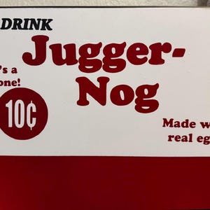 May include: A white and red metal sign featuring the text DRINK Jugger-Nog in large red letters. It includes the phrases That's a cold one! and Made with real eggs! along with a red circle containing the text 10 cents in white font.