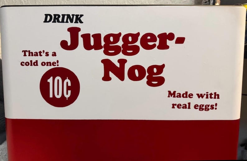 May include: A white and red metal sign featuring the text DRINK Jugger-Nog in large red letters. It includes the phrases That's a cold one! and Made with real eggs! along with a red circle containing the text 10 cents in white font.