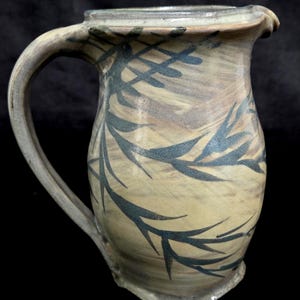 May include: A ceramic pitcher with a beige and tan marbled glaze finish, featuring a dark charcoal-colored painted leaf pattern that wraps around the body. The pitcher has a curved handle on the left side and a small pouring spout at the top rim.