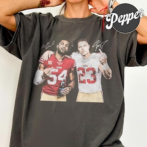 Könnte beinhalten: Ein anthrazitfarbenes Kurzarm-T-Shirt mit einem Grafikdruck von zwei Footballspielern in 49ers-Trikots mit den Nummern 54 und 23 sowie Unterschriften darüber. Der Spieler im roten Trikot hält eine Flasche, beide Spieler gestikulieren.