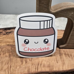Puede incluir: Una pegatina con un tarro de crema de chocolate de dibujos animados con una cara sonriente y la palabra "Chocolate" en rojo. El tarro es marrón con una tapa y borde blancos, sobre un fondo de madera.