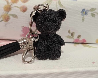 May include: A black teddy bear keychain with a silver key ring and a black tassel. The bear is made of a textured material and has a cute, friendly expression.