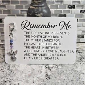 May include: A white card with the text "Remember Me" and a message about the meaning of the stones, heart, and angel charm attached to the card. The charm is silver and has a purple and blue crystal.