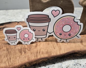 May include: Three cartoon stickers of a coffee cup, a doughnut, and a heart. The coffee cup and doughnut are holding hands. The doughnut is pink with pink sprinkles and a bite taken out of it. The coffee cup is brown with a white lid. The heart is pink.