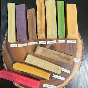May include: A collection of rectangular dog chews in various colors including yellow, purple, brown, green, and red. Each chew is labeled with white paper tags reading Apple, Blueberry, Blackberry, Smoked, Coconut, Mint, Pumpkin, PB, Bacon, Reg Cheese, and Strawberry.