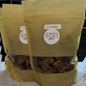 May include: Several bags of pet treats are displayed. Each bag is a light brown color and contains brown, cube-shaped treats. The bags have a white label that reads "8 oz Rabbit" and "MY PET shop".