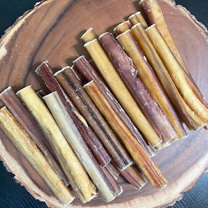 May include: A collection of dog chews in various shades of brown and tan, arranged on a round wooden serving board. The chews have a cylindrical shape and a textured surface, with some showing a reddish hue.