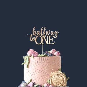 May include: Gold glitter cake topper with the text "halfway to ONE" on a pink cake decorated with flowers.