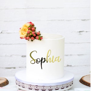 May include: White cake with a gold glitter 'Sophia' topper and a single yellow rose with red berries.