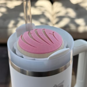 May include: A white Stanley mug with a pink and tan concha-shaped lid and a clear straw. The mug has a white handle and a silver band near the top. The Stanley logo is visible on the front of the mug.